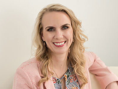 A woman with long, wavy blonde hair is smiling, wearing a light pink blazer and a floral-patterned top, sitting against a light-colored background, ready to deliver her commencement speech for an online college. Saint Joseph's College of Maine