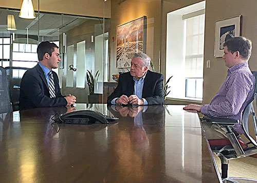 Three men in business attire are seated at a large conference table having a conversation in an office setting, discussing Governor Brennan's legacy. Saint Joseph's College of Maine