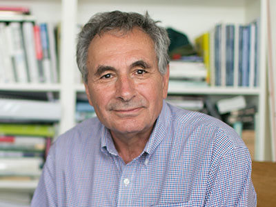 A man with gray hair and a plaid shirt is sitting in front of a bookshelf filled with books and binders. He is looking directly at the camera, evoking Ron Phillips' dedication to Community Economic Development. Saint Joseph's College of Maine