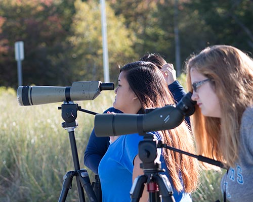 Two women, embodying a passion for science, observe through telescopes on tripods outdoors, with trees swaying gently in the background. One woman is in a blue shirt; the other in a grey shirt and sunglasses. Saint Joseph's College of Maine