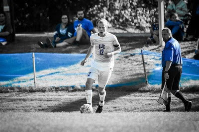 A soccer player in a white jersey with the number 12 dribbles the ball on a pristine turf field. Spectators and an official are visible in the background, adding to the competitive edge of the game. Saint Joseph's College of Maine