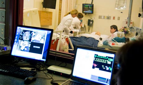 Medical professionals attending to a patient in a high-tech hospital room at Mercy Hall, while monitors display vital signs; view includes a computer setup in a separate monitoring area. Saint Joseph's College of Maine