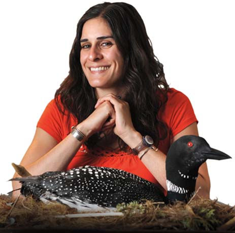 A woman with long dark hair, wearing a red shirt, smiles and sits behind a taxidermy loon on a nest—a scene that captures why people love loons. Saint Joseph's College of Maine