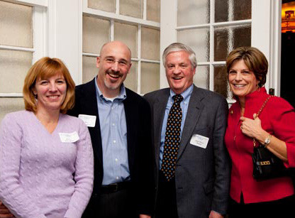 Four adults stand together smiling, dressed in business casual attire with nametags on their chests. They are indoors near paned glass doors, preparing for the President's Society Dinner. Saint Joseph's College of Maine
