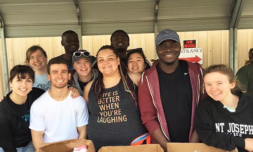 A group of smiling people stand under a shelter, some wearing casual clothing and one with a shirt that reads "Life is better when you're laughing." They appear to be students on a Spring Break service trip, volunteering at an event. Saint Joseph's College of Maine