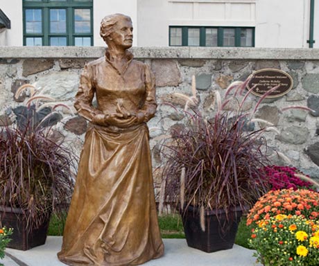 Bronze sculpture of a woman, possibly a founder of the Sisters of Mercy, standing with hands clasped in front, placed outdoors between two potted plants, in front of a stone wall with a plaque. Saint Joseph's College of Maine