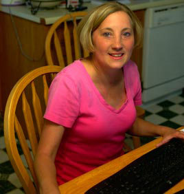 A woman in a pink shirt sits at a table with a keyboard in front of her. Fresh from a successful lung surgery, she is smiling and looking at the camera. Saint Joseph's College of Maine
