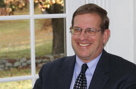 A man with glasses in a suit and tie smiles while sitting indoors near a window showcasing a yard and trees. Engrossed in good reads, he embodies the spirit of good minds, finding both inspiration and tranquility in his surroundings. Saint Joseph's College of Maine