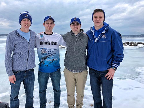 Four young men standing on a frozen lake, wearing casual winter clothing, including jackets, hats, and jeans, with a cloudy sky and rocky shoreline in the background. They seem to embrace their wild side as they prepare for an adventurous day of ice fishing. Saint Joseph's College of Maine