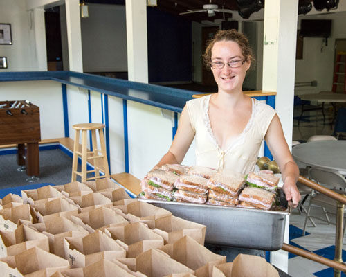A person standing indoors, smiling and holding a tray filled with pre-packaged sandwiches, next to a table with multiple paper bags, embodies the spirit of our Summer Meals Program, supporting local communities. Saint Joseph's College of Maine