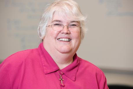 A person with short white hair wearing glasses and a pink top smiles at the camera, reminiscent of Dr. Susan Miovech, a revered 1982 alumni. Saint Joseph's College of Maine