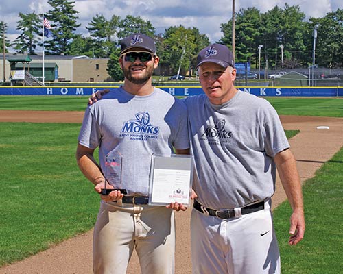 Two men in matching shirts and baseball caps stand on a baseball field holding an award and a certificate. The man on the left is smiling with his trophy, while the other has his arm around him. It looks like their hard work both on the field and hitting the books has truly paid off. Saint Joseph's College of Maine