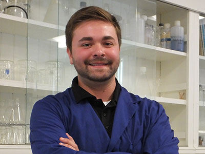 A man wearing a blue lab coat stands with his arms crossed in front of glass shelves filled with laboratory glassware, looking every bit the part of a Saint Joseph’s College Commencement speaker. Saint Joseph's College of Maine