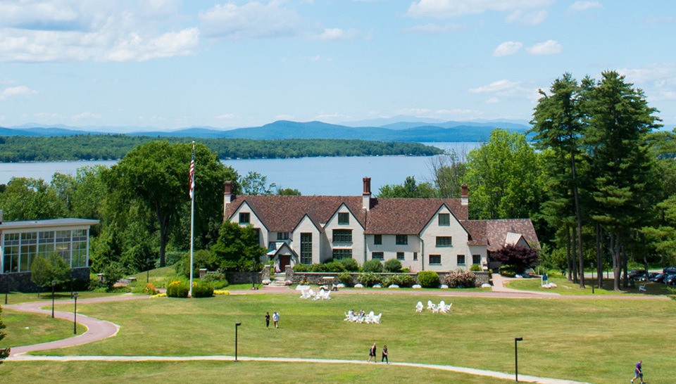 A large white house with a red roof sits near a lake, surrounded by green lawns, trees, and distant mountains under a partly cloudy sky. Saint Joseph's College of Maine
