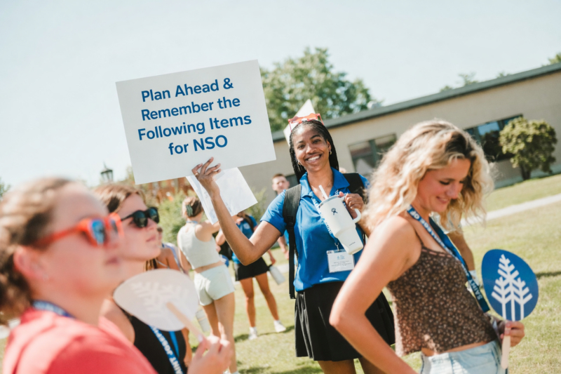 A smiling person holds a sign reading “Plan Ahead & Remember the Following Items for NSO” while standing outdoors among a group of students on a sunny day. Saint Joseph's College of Maine