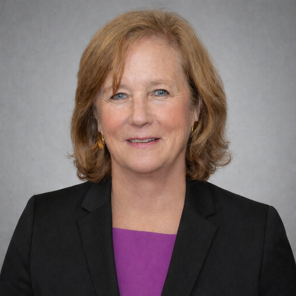 A woman with shoulder-length light brown hair wearing a black blazer over a purple top, posing and smiling as if ready for a graduation ceremony, stands in front of a plain gray background. Saint Joseph's College of Maine