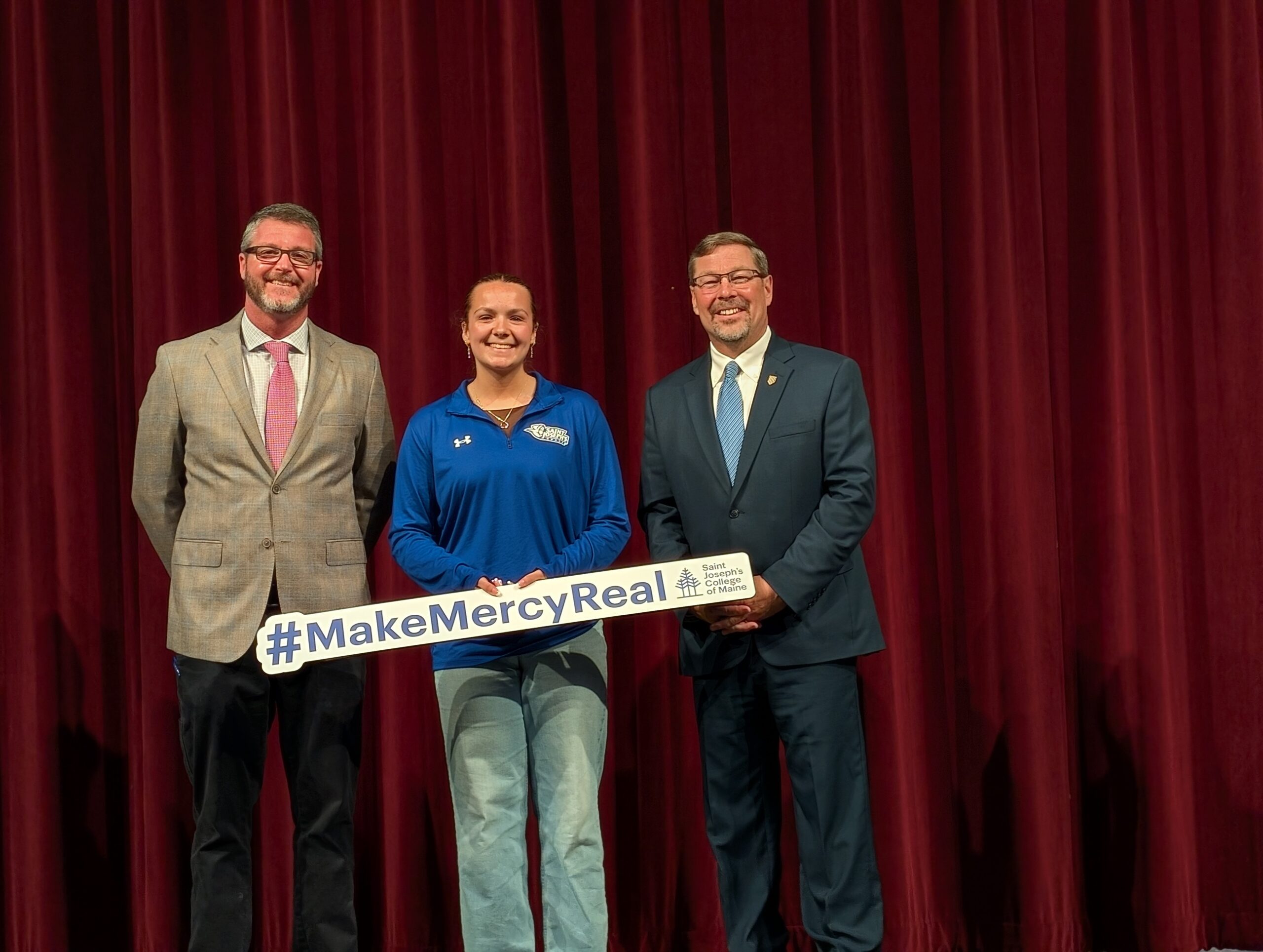 Three people stand smiling in front of a red curtain. Adriana Pettengill, in the center wearing a blue athletic pullover, holds a #MakeMercyReal sign. The two beside her wear suits, celebrating her full ride achievement. Saint Joseph's College of Maine