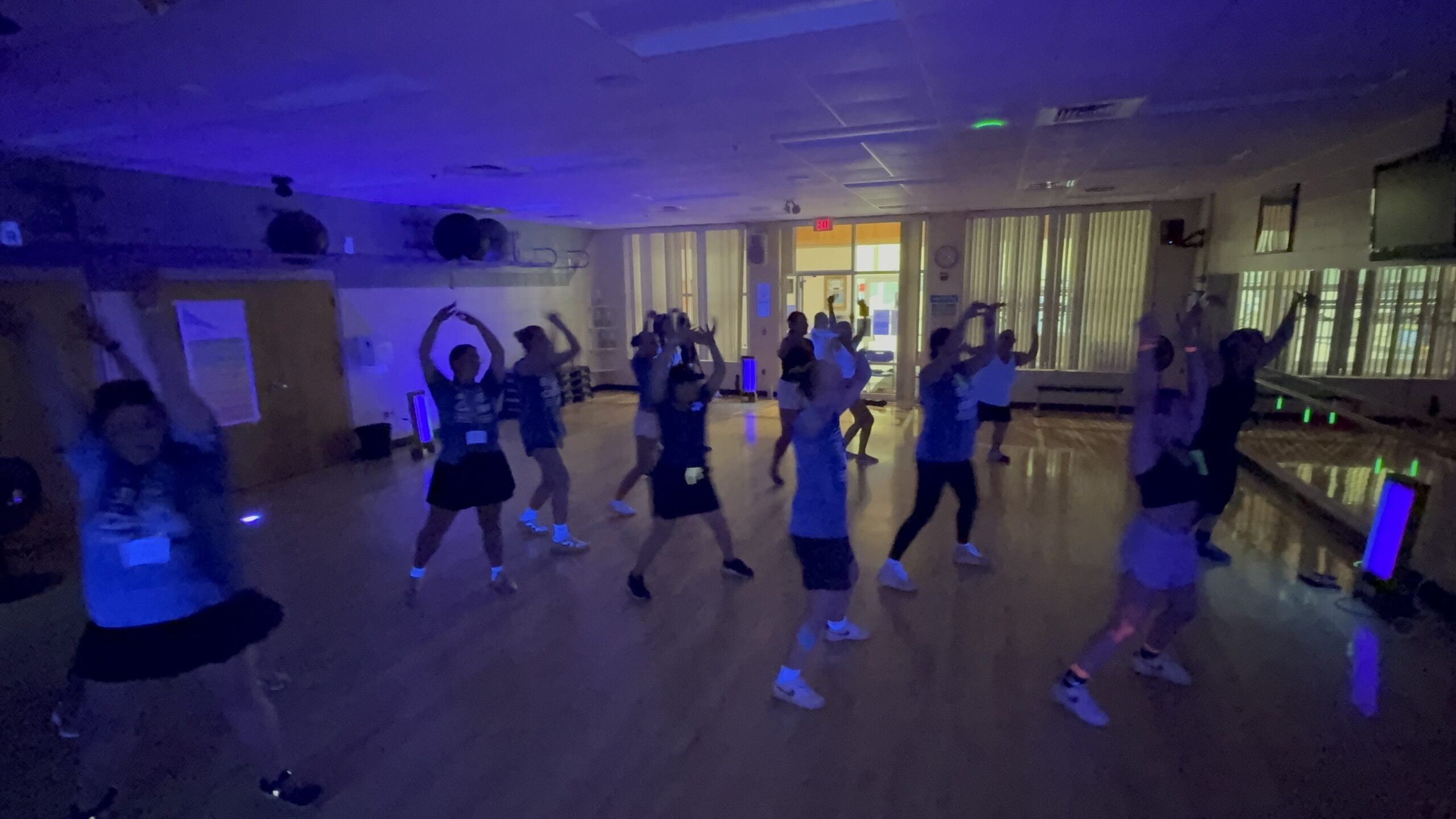 A group of people dance together in a dimly lit fitness studio with purple and blue lighting, raising their arms and moving energetically—perfect for employee wellness programs. Mirrors line one wall, and exercise equipment is visible in the background. Saint Joseph's College of Maine