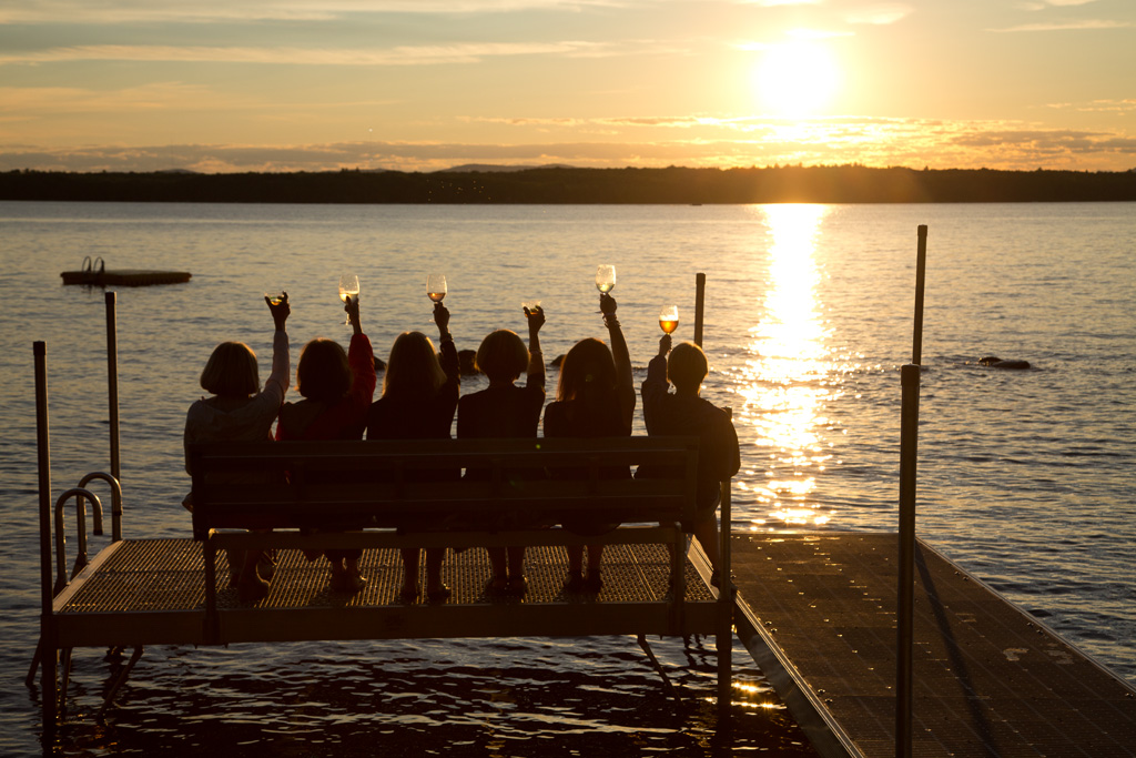 Six people sit on a dock bench by a lake at sunset, raising glasses in celebration, with golden sunlight reflecting on the water. Saint Joseph's College of Maine