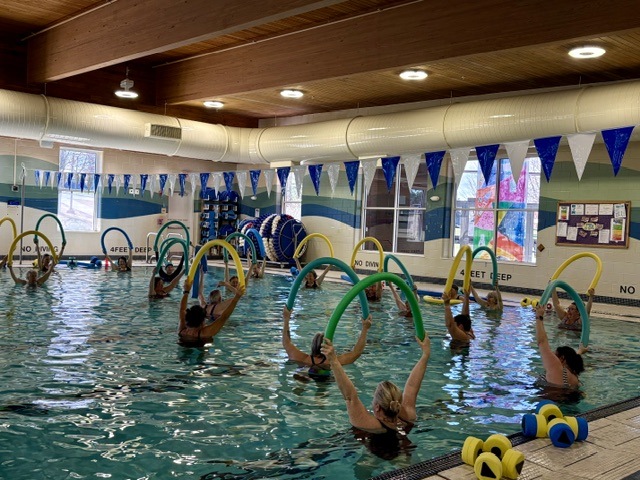 A group of people in a swimming pool participate in an aquatic fitness class, each holding colorful foam pool noodles above their heads. Blue and white flags hang overhead, and exercise equipment is visible on the pool deck. Saint Joseph's College of Maine