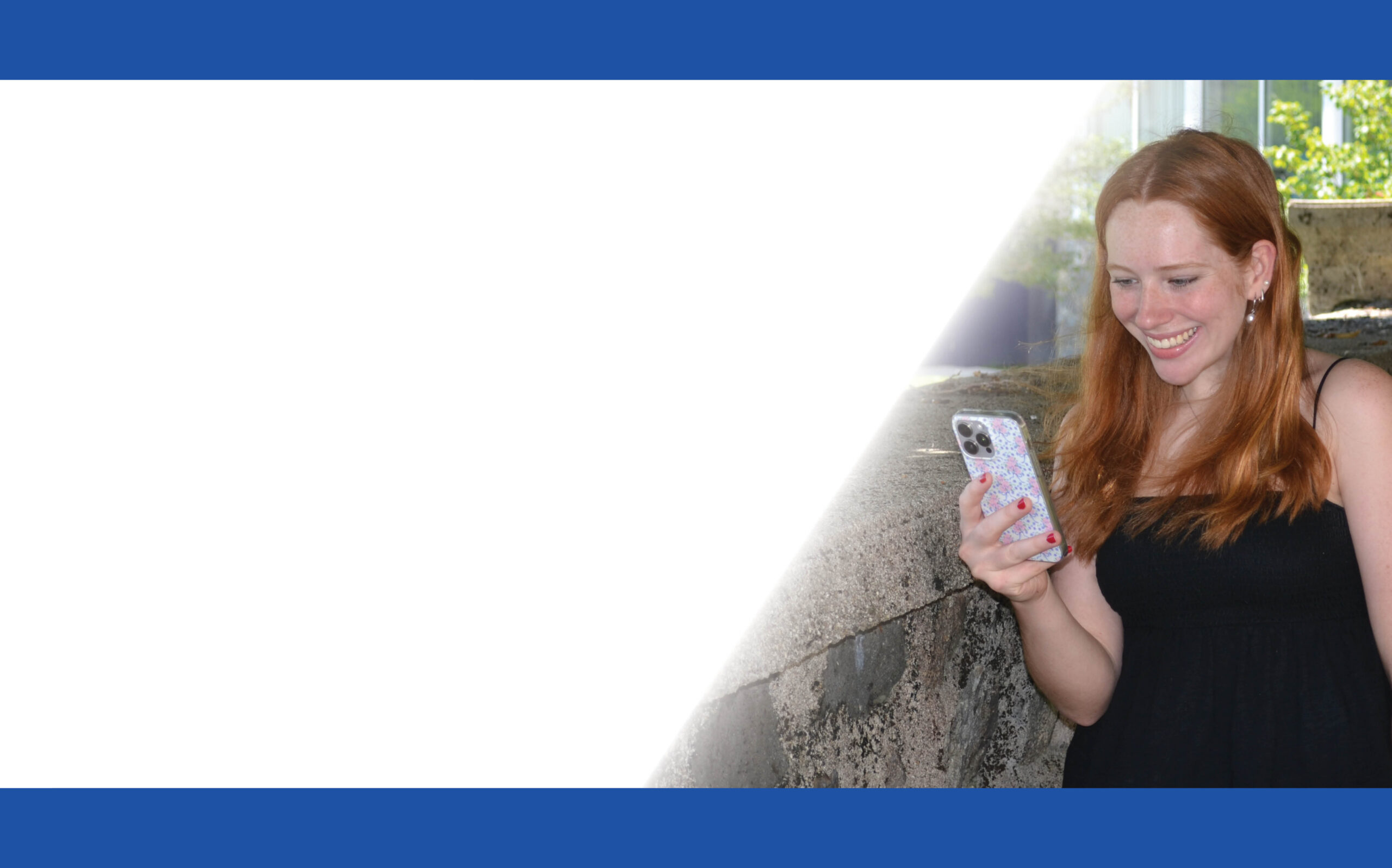 A woman with long red hair in a black dress smiles while looking at her phone. She stands outside near a stone wall on the campus of Saint Joseph’s College of Maine. The left side of the image has a white space bordered by blue bars. Saint Joseph's College of Maine
