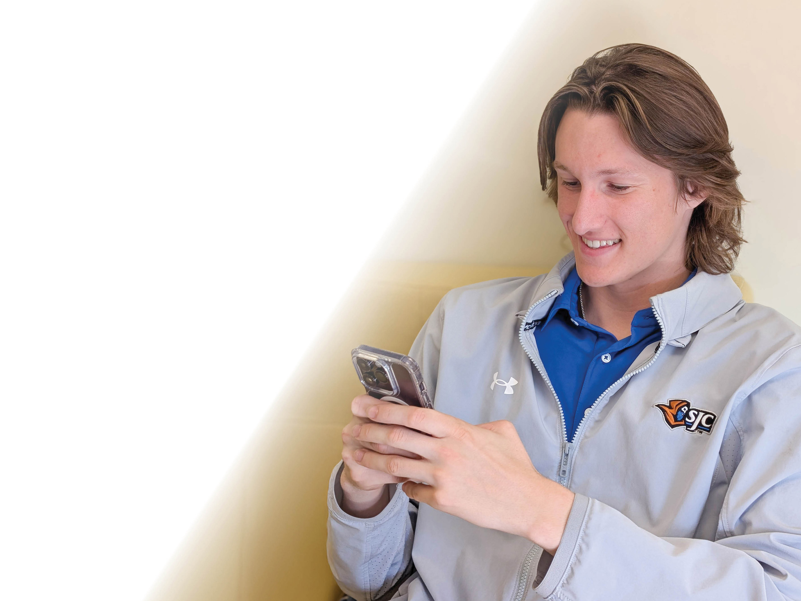 A young person with long hair in a gray Saint Joseph’s College of Maine (SJC) jacket smiles while looking at their phone, sitting on a light-colored chair against a bright background. Saint Joseph's College of Maine