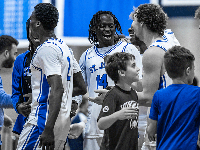 Basketball players in white jerseys from St. Joseph's College celebrate on the court while children and others in blue shirts stand nearby. One player wearing number 14 is smiling in the center, capturing a moment of joy reminiscent of the spirited games seen at Saint Joseph’s College of Maine. Saint Joseph's College of Maine