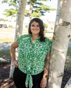 A person with long dark hair, wearing a green and white patterned shirt and black pants, stands smiling outdoors next to a white tree trunk on a sunny day, embodying the confidence gained through Career Studio’s career development support. Saint Joseph's College of Maine