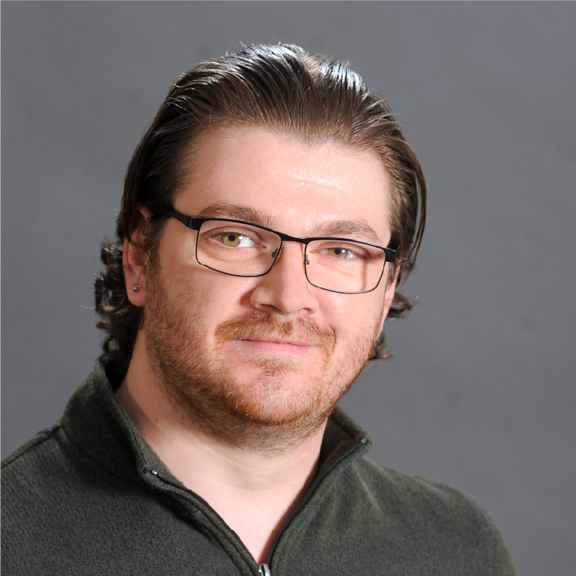 A man with light skin, brown hair, and glasses is wearing a green zip-up sweater and smiling slightly. The background is plain gray, and he closely resembles Gregory Wadsworth. Saint Joseph's College of Maine