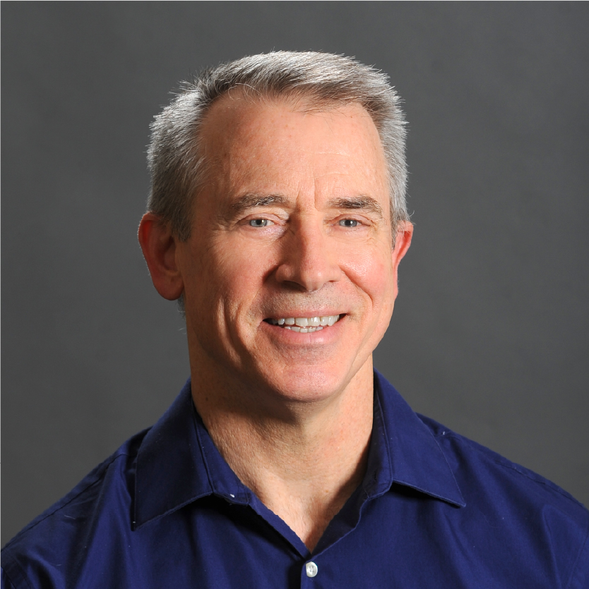 A man with short gray hair wearing a blue collared shirt smiles in front of a plain gray background. Saint Joseph's College of Maine