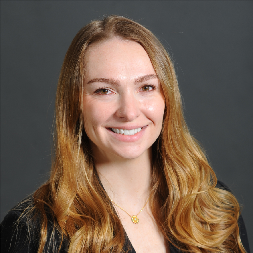 A woman with long, wavy blonde hair smiles at the camera. She is wearing a black top and a gold necklace, with a neutral gray background. Saint Joseph's College of Maine