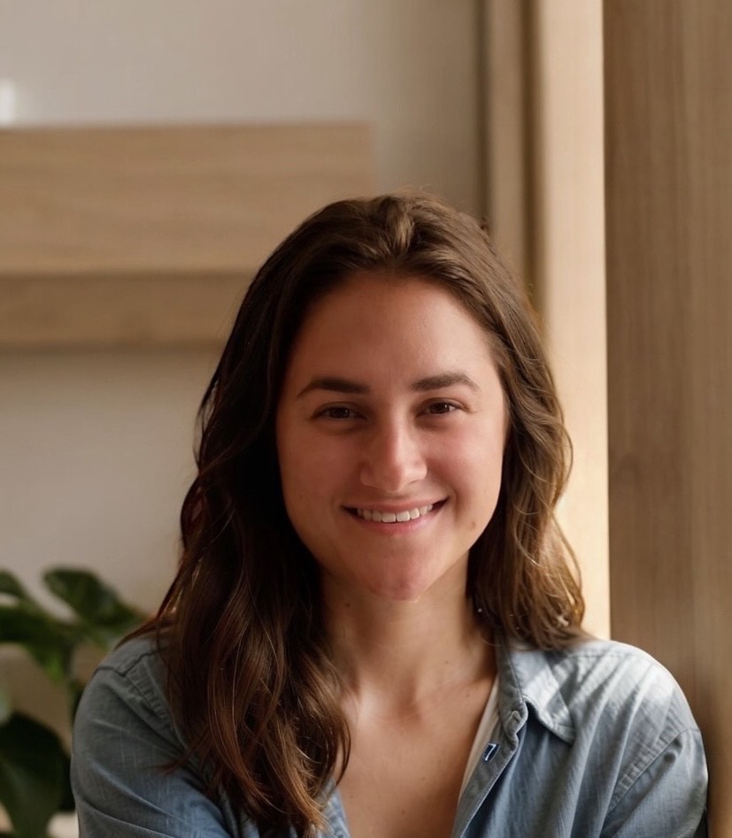 A young woman with long brown hair, wearing a light blue shirt, smiles warmly while sitting indoors with soft natural light and a plant in the background. Kiley Breitenfeld, MSN, RN, exudes a sense of calm professionalism in this inviting setting. Saint Joseph's College of Maine