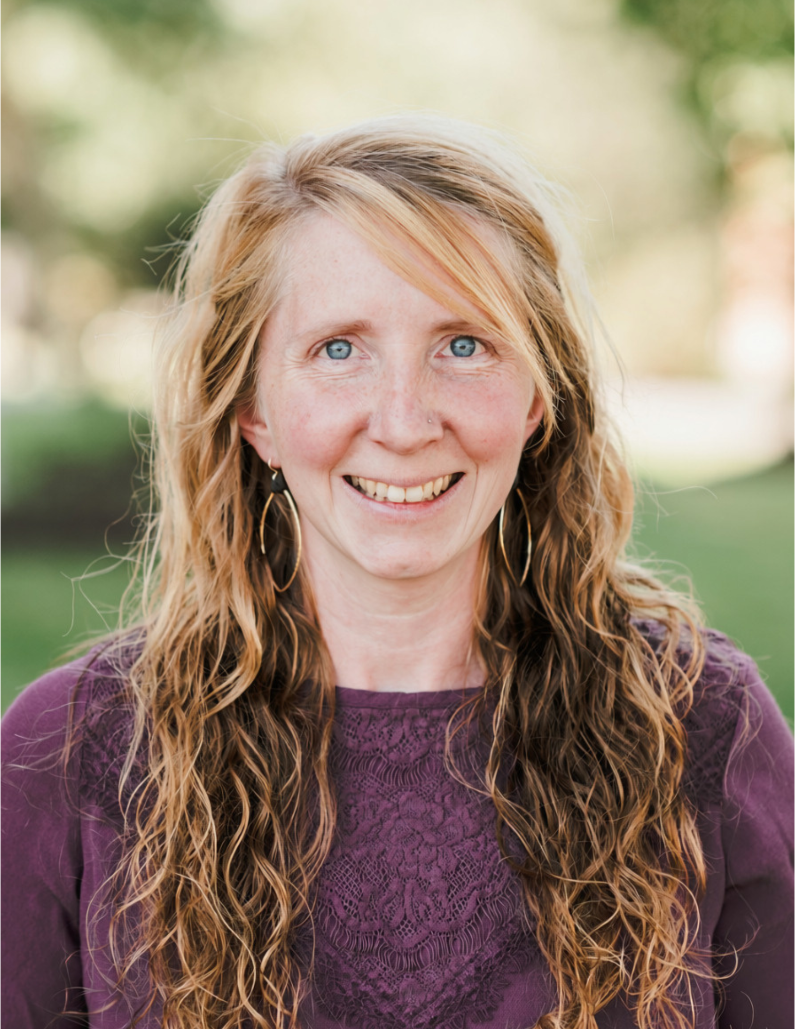 Woman with wavy, long blonde hair wearing a purple top and large hoop earrings, smiling outdoors with a blurred green background. Saint Joseph's College of Maine