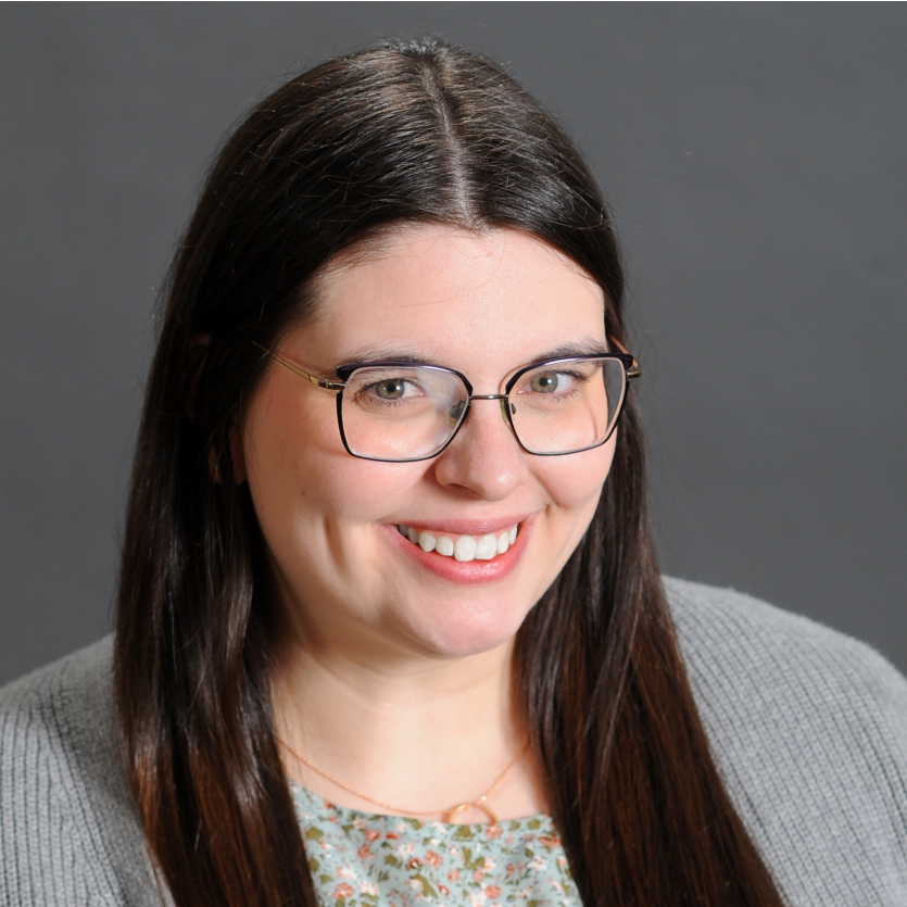 A woman with long brown hair and glasses smiles at the camera. She is wearing a gray sweater over a floral top and sits in front of a plain dark gray background. Saint Joseph's College of Maine