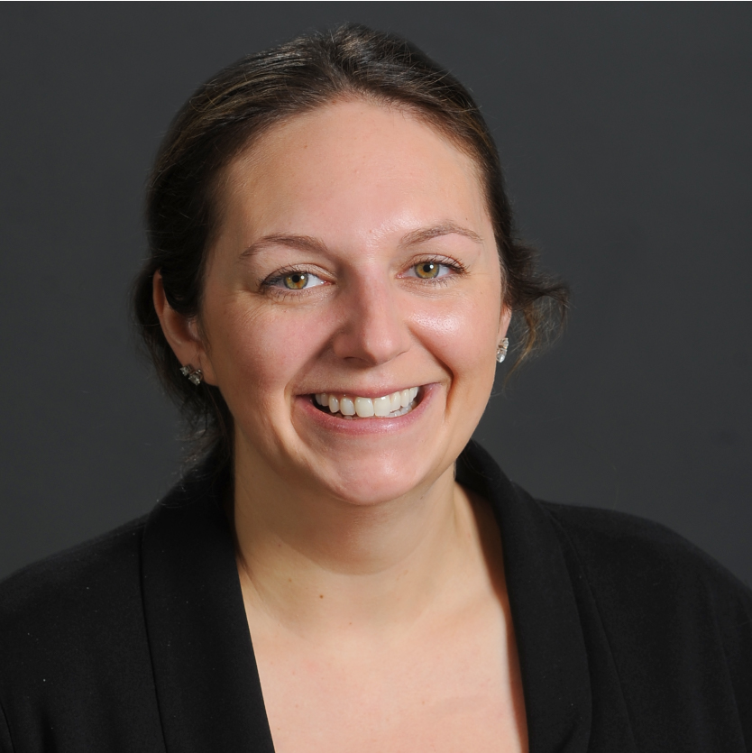 A woman with brown hair, tied back, smiles at the camera. She is wearing stud earrings and a black top. The background is plain and dark gray. Saint Joseph's College of Maine