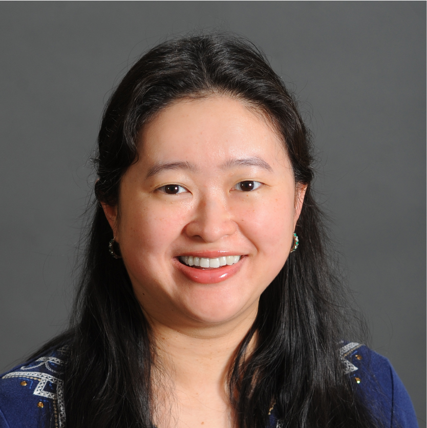 A woman with long black hair smiles at the camera. She is wearing a blue top with white patterns and small earrings. The background is plain gray. Saint Joseph's College of Maine