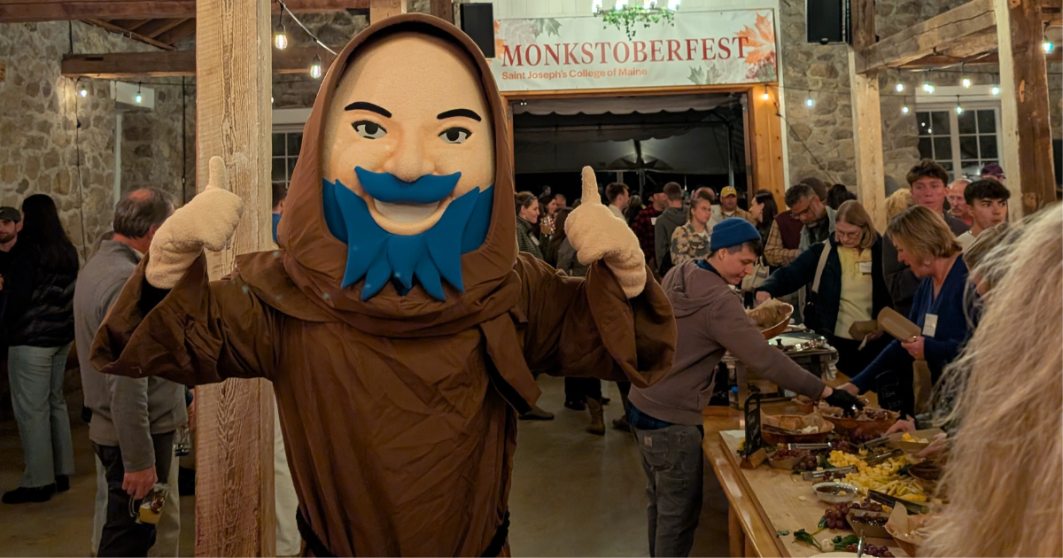 A person in a monk mascot costume with a blue beard poses, smiling and giving thumbs up at Saint Joseph’s College of Maine’s MONKSTOBERFEST, where people are gathered around a table of food. Saint Joseph's College of Maine