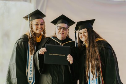 Three graduates in caps and gowns smile while holding an Integrated Studies diploma cover from Saint Joseph’s College. Saint Joseph's College of Maine