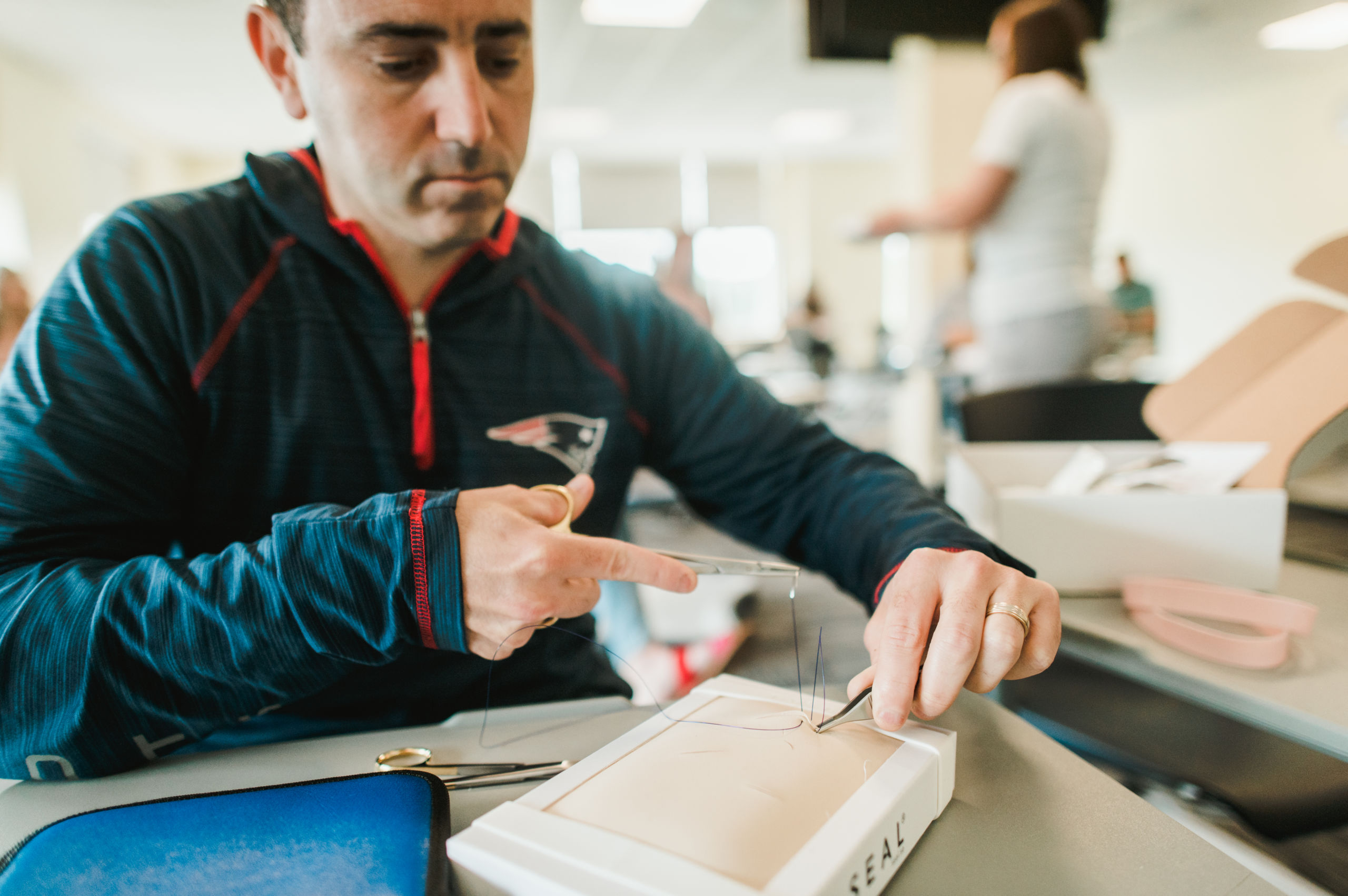 2_Nursing_June2021_032_Hr A man in a dark jacket practices suturing on a rectangular silicone model at a table, using a needle holder and other surgical instruments. Saint Joseph's College of Maine