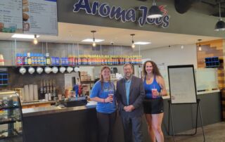 Three people stand in front of an Aroma Joe's coffee shop counter at the bustling SJC campus. Two women hold drinks while a man in a suit stands between them. A menu and various counter items are visible in the background, capturing the lively atmosphere. Saint Joseph's College of Maine
