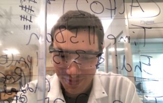 A person wearing glasses and a lab coat is looking intently through a glass board covered with various handwritten notes and equations. Saint Joseph's College of Maine