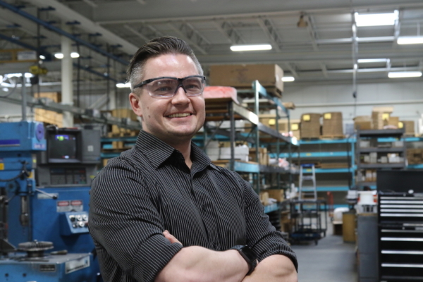 Bryon Williams poses, wearing safety glasses, on the work floor in front of shelves of tools