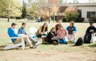 A group of people sit on Adirondack chairs and on the grass in a park-like setting with buildings and trees in the background. Some are looking at a laptop, likely discussing their achievements from the Fall 2019 semester, perhaps even celebrating those who made the Dean’s List. Saint Joseph's College of Maine