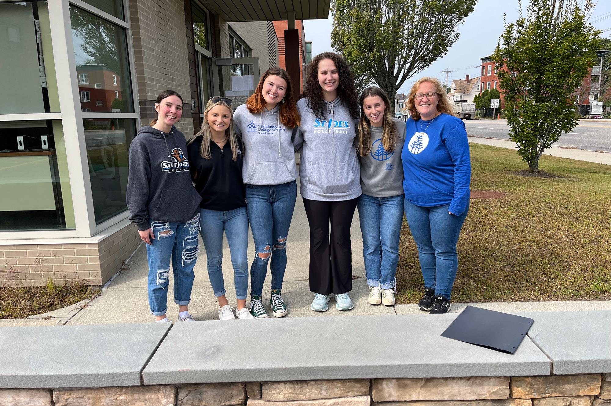 Six women are standing in a line outdoors, smiling at the camera. They are casually dressed, wearing jeans and a mix of sweatshirts and hoodies. They're next to a building with a sidewalk and grassy area, possibly having just finished some Community Service at Mercy Center. Saint Joseph's College of Maine
