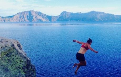 A person in a red top and black shorts jumps off a cliff into a deep blue body of water with mountains in the background under a clear sky, experiencing pure trail magic. Saint Joseph's College of Maine
