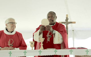 Two clergy members in red vestments stand at an altar; the central figure holds up a large wafer, and religious items are positioned in front of them, including a chalice and a crucifix. The scene embodies the tradition of compassionate action, reflecting mercy through their sacred rituals. Saint Joseph's College of Maine