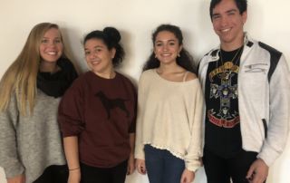 Four people standing side by side, facing the camera, against a plain white background. They are casually dressed and smiling. Saint Joseph's College of Maine
