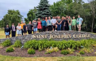 A group of individuals stand together behind a stone sign that reads "Saint Joseph’s College of Maine" in a grassy, tree-lined area, possibly discussing New England Gothic Literature. Saint Joseph's College of Maine
