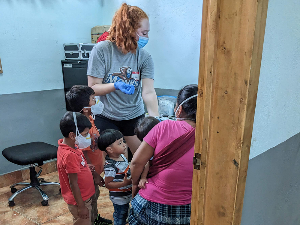 A woman wearing a mask and gloves attends to a group of children and an adult in a small room with a chair and medical supplies. All individuals are masked, and the woman wears a grey t-shirt, exemplifying the dedication often seen during international service trips. Saint Joseph's College of Maine