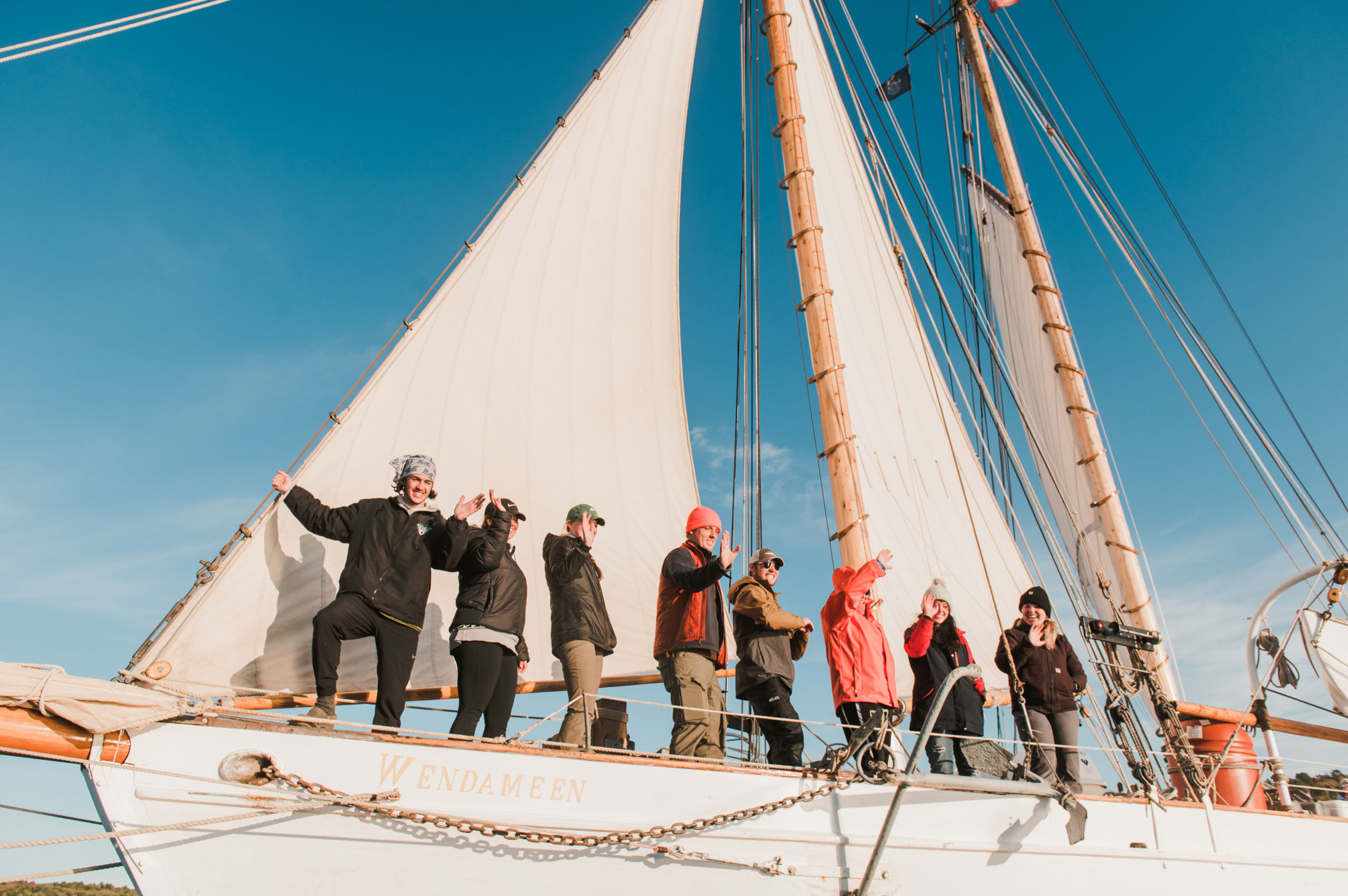 Learnherethere-Scaled A group of people stand on the deck of a sailboat named WENDAMEEN, beneath the raised sails, on a sunny day. Some are waving and smiling while others look out to the horizon. Saint Joseph's College of Maine